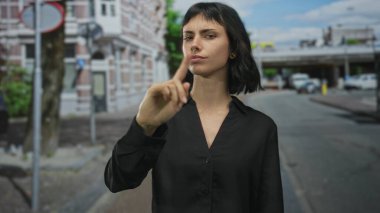 Siyah gömlekli kadın, tuğla bina cephesi ve park halindeki arabalarla sokakta parmak ucuyla işaret ediyor; inkar.