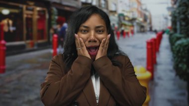 Bir kaldırım taşı şehir caddesinde kahverengi bir palto giyerken yanaklara ellerle bastıran kadın kırmızı kalın çizgili; sürpriz sevinç şaşkınlığı.