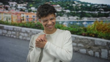 Gözlüklü ve beyaz kazaklı bir adam, taştan bir duvar ve uzak bir yamaç köyü ile güneşli bir sahil geçidinde el sıkışıyor..