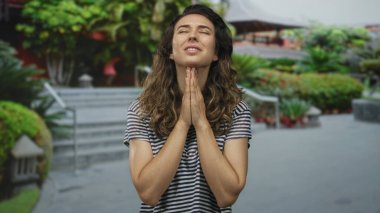 Çizgili tişörtlü genç İspanyol kadın başını yukarı eğerken avuçlarını göğsüne yapıştırıyor. Çevresinde yeşillik, umut var..