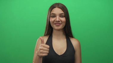 Genç beyaz kız parmak göğse işaret ediyor ve siyah askılı bluz giyerken stüdyoda yeşil arka planla baş parmak işareti yapıyor..