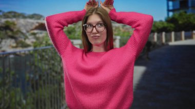 Pembe kazaklı ve gözlüklü kadın güneş ışığı altında gelişigüzel kollarını başının arkasına kaldırıyor..