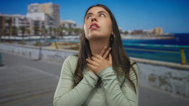 Deniz kenarındaki güvertede boğazı sıkışan, etrafı okyanus ve şehir zeminiyle çevrili genç İspanyol kadın, açık hava ortamında gerilimin altını çiziyor..