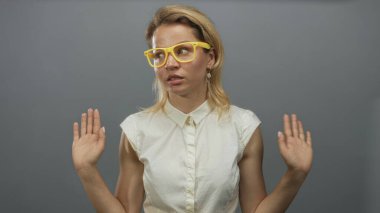 Sarı gözlüklü ve beyaz gömlekli sarışın kadın stüdyo ortamında omuz silkme hareketinde iki elini de kaldırıyor..
