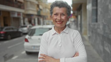 Gri saçlı ve çapraz kollu bir kadın şehir sokağında arka planda arabalarla samimi bir şekilde gülümsüyor, beyaz giyinmiş, neşeli ve dostane bir açık hava sahnesini vurguluyor..