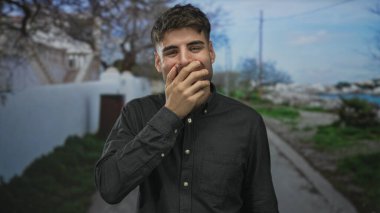 İspanyol genç adam gülümseyen ağzı güneş ışığı altındaki sokakta beyaz duvar ve ağaçlarla kaplıyor; kahkaha neşe neşesi.