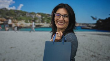 Güneşli sahil şeridinde el kaldırarak alışveriş çantası tutan gülümseyen kadın, mutluluk rahatlaması..