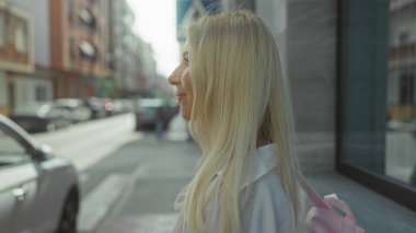 Park halindeki arabaların ve vitrinlerin yanında pembe sırt çantalı ve uzun sarı saçlı bir kadın yürüyor..