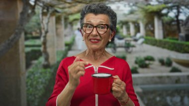 Gri saçlı, kırmızı kazaklı bir kadın yeşil yeşil bir parkta kahve fincanı tutuyor, ağaçlar ve bitkilerle çevrili, dingin ve açık hava manzarası sunuyor..