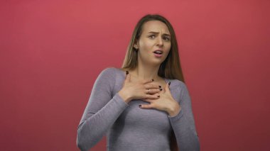 Kadın, elleri göğsüne bastırılmış bir şekilde izole edilmiş kırmızı bir arka plana karşı korkusunu dile getiriyor, genç beyaz bir kadının duygularını ve tepkisini canlı bir şekilde gösteriyor..