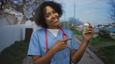 Mavi önlük içinde steteskop takan kadın doktor sokaklarda parmak ucuyla ilaç şişesini işaret ediyor; şefkat ve sağlık güvencesi.