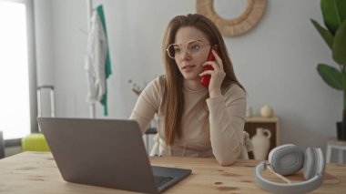 Binadaki ahşap bir masada dizüstü bilgisayara bakarken kırmızı akıllı telefonu kulağına tutan kadın. Konsantrasyon..
