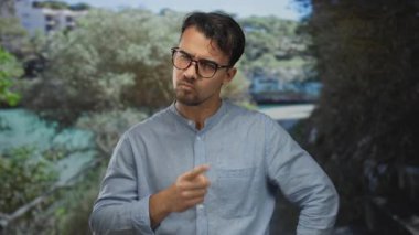 Genç adam park ortamında hayal kırıklığı yaşıyor, gözlük takıyor ve açık havada dinamik yeşil bir sahne altında mavi bir gömlek giyiyor..