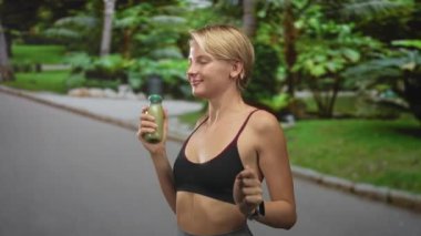Sokak yolunda elinde yeşil meyve suyu şişesi tutan sarışın bir kadın, kollarını kaldırmış ve rahat bir şekilde koşarken gülümsüyor; sağlık keyfi.