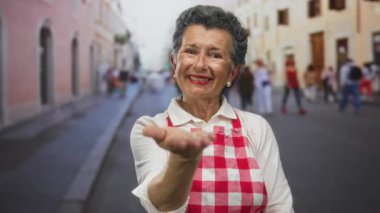 Gri saçlı ve önlüklü yaşlı bir kadın hareketli bir eski kasaba caddesinde sıcacık gülümsüyor. Bulanık yayalar ve tarihi mimariyle şehir ortamını sergiliyor..