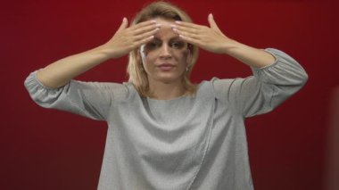 Sarı saçlı, gri bluzlu bir kadın iki elini havaya kaldırıyor ve kırmızı stüdyo alanında genişleyen gözler. Sürpriz..