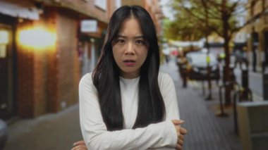 Genç Çinli bir kadın hareketli bir caddede, şehir manzarası ve arka planda hafif ışıklandırmayla çevrili, şaşkın bir ifadeyle ayakta duruyor..