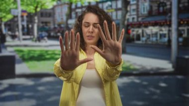Sarı gömlekli kadın, elleri havada, avuç içi açık, şehir caddesinde dur hareketi yapıyor; korku alarmı tepkisi.
