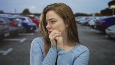 Bir sokak otoparkında arabaların önünde ellerini çenesine yapıştırmış genç bir kadın. Endişe belirsizliği..