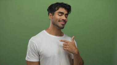 Gözleri kapalı beyaz tişört giyen ve yeşil stüdyoda göğsünü gösteren parmak gösteren İspanyol asıllı genç adam..