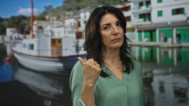 Yeşil bluzlu kadın, liman binasına yanaşmış beyaz teknenin yanında baş parmak hareketi gösteriyor..