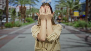 Gözlüklü, bej gömlekli bir kadın, güneşli caddede palmiye ağaçlarıyla kaplı gözlerini kapatıyor..