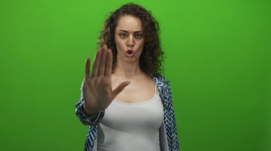 Avuç içleri açık genç bir İspanyol kadın yeşil stüdyoda, beyaz atlet bluz ve desenli gömlek giyerek durmak için el hareketi yapıyor..