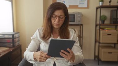 Ofis binasında elleri ekranda bağlı bir şekilde tablet okuyan bir kadın; iş stresi ve konsantrasyon.
