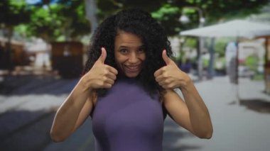 Güneşli bir günde güneşli bir sokakta neşeli bir gülümsemeyle baş parmak kaldıran genç bir kadın kendine güvenen bir ifadeyle pozitif titreşimler sergiliyor..