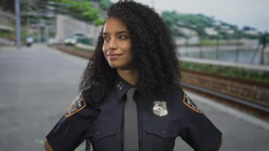 Üniformalı kadın polis memuru, açık hava tren istasyonunda kendine güvenerek duruyor. Hafif bir gülümsemesi ve kıvırcık saçlarıyla..
