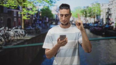 Akıllı telefon tutan ve parkedilmiş bisikletli bir sokak kanalı köprüsünü işaret eden adam, bir mesajı kontrol ederken mavi beyaz çizgili tişört giyiyor; tefekkür.