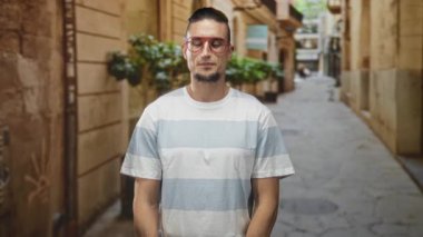 Sessiz kalmak için parmak parmağa duran adam, kırmızı gözlüklü ve dar bir sokakta açık çizgili gömlek giyen, nötr bakış ve rahat duruş; sessiz gizlilik.