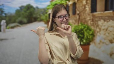 Gözlüklü sarışın kadın, antik taş binanın önünde kendini işaret ediyor..