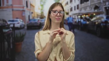 Bej gömlekli bir kadın, park halindeki araba ve binalarla dolu bir kaldırım taşı caddesinde ellerini göğsüne yaklaştırıyor..