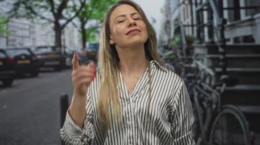 Sarışın, orta yaşlı, uzun saçlı, çizgili tişörtlü, yeşil caddede parmağıyla başını gösteren bir kadın..