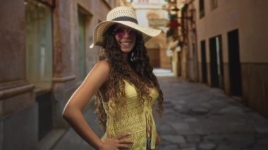 Geniş hasır şapka ve mor güneş gözlüğü takan kadın, dar bir kaldırım taşı sokakta elleri kalçalarında gülümsüyor; neşe yolculuğu.