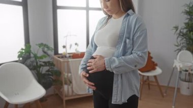 Hamile kadın huzurlu bir bebek odasında duruyor, elleri karnının üstünde, rahat bir ev dekoruyla çevrili, beklenti ve sıcaklığı yansıtıyor..