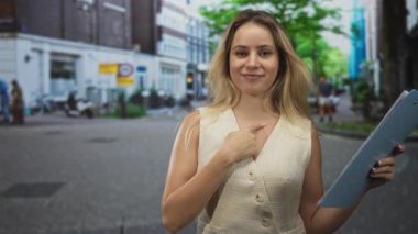 Genç sarışın kadın, scooter ve tuğla binaların yanındaki sokakta elinde pano tutarken parmak göğse işaret ediyor..