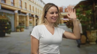 Kadın, dükkanlar ve saksılarla dolu bir yaya caddesinde el sallıyor ve gülümsüyor. Karşılama jesti olarak ileriye doğru yürüyor..