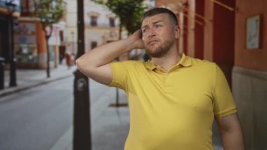 Sokak sokak lamba direği ve mağaza vitrinine bakarken, sarı polo tişört giyen bir adam yanağına elle dokunuyor..