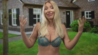 Sarışın bir kadın kollarını silkiyor ve binanın önündeki gümüş dantelli sütyenle göğüs dekoltelerini gösteriyor..