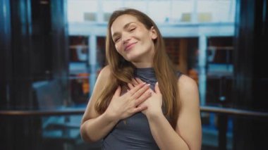 Bir kadın, iki elini de bir tekne güvertesine koyup gülümsüyor modern cam bina cephesinin önünde tırabzanlar açıyor; minnettarlık..