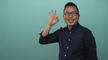 Turkuaz stüdyoda gözlüklü ve koyu renk gömlekli bir adam el hareketi yapıyor ve gülümsüyor..