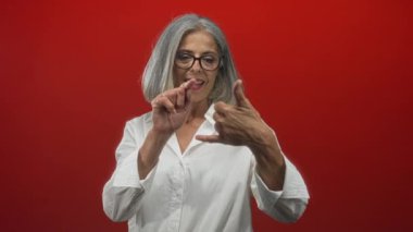 Beyaz gömlekli, gri saçlı ve gözlüklü bir kadın, stüdyoda kırmızı duvara parmağını uzatırken parmaklarını sayıyor..