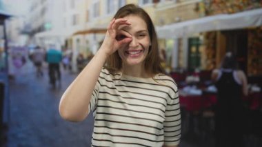 Çizgili tişörtlü kadın güneşli bir restoranın terasında eli gözünün önünde işaret veriyor..