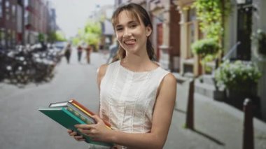 Kitap istiflemiş, dili dışarıda gezen ve kalabalık bir caddede bisiklet ve saksılarla gülümseyen bir kadın; oyuncu öğrenci iyimserliği.
