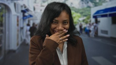 Kahverengi ceketli kadın, kalabalık bir şehir caddesinde yoldan geçenlerin ortasında elleri ile ağzını kapatır; neşeli sürpriz.