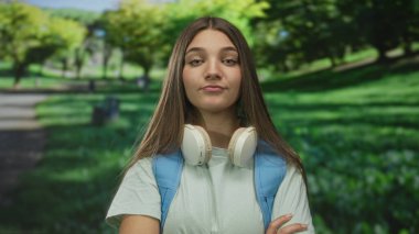 Beyaz kulaklıklı ve mavi sırt çantalı bir kız. Kavis yolunun yakınındaki güneşli ormanda kollarını kavuşturuyor..