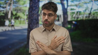 Uzun çam ağaçlarının altında yeşil parkta kollarını bağlayan bej gömlekli bir adam. Hayal kırıklığını inkar ediyor..