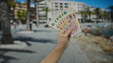 Arka planda bulanık sahil manzaralı Arjantin pesoları tutan adam, paranın dinamik havasını yakalıyor ve deniz kenarı kentlerinde seyahat ediyor..